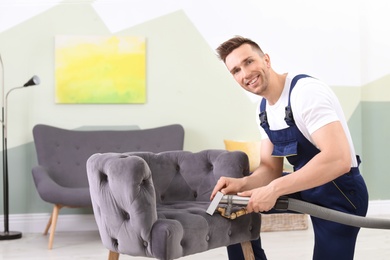 Male worker removing dirt from armchair with professional vacuum cleaner indoors