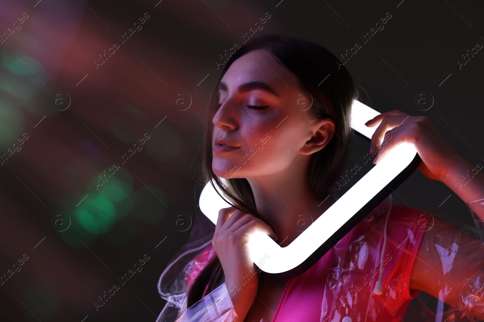 Photo of Fashionable portrait of beautiful woman wearing transparent coat with square lamp on dark background in neon lights. Space for text
