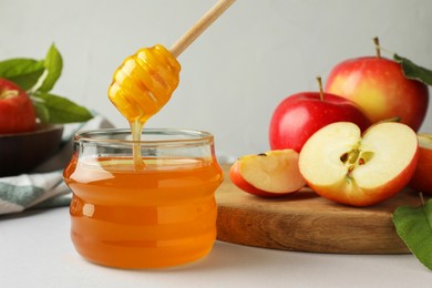 Photo of Dripping sweet honey from dipper into jar and fresh apples on white table