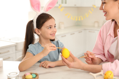 Photo of Happy daughter with bunny ears headband and her mother painting Easter egg in kitchen