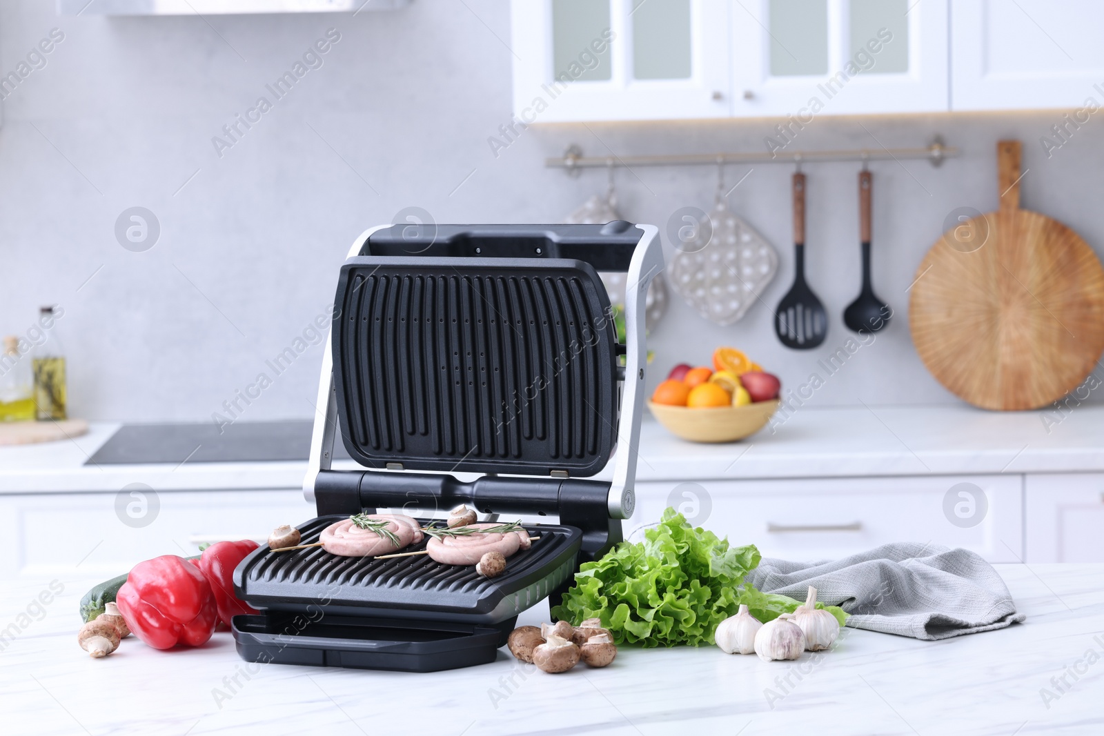 Photo of Electric grill with homemade sausages, rosemary and vegetables on white table in kitchen