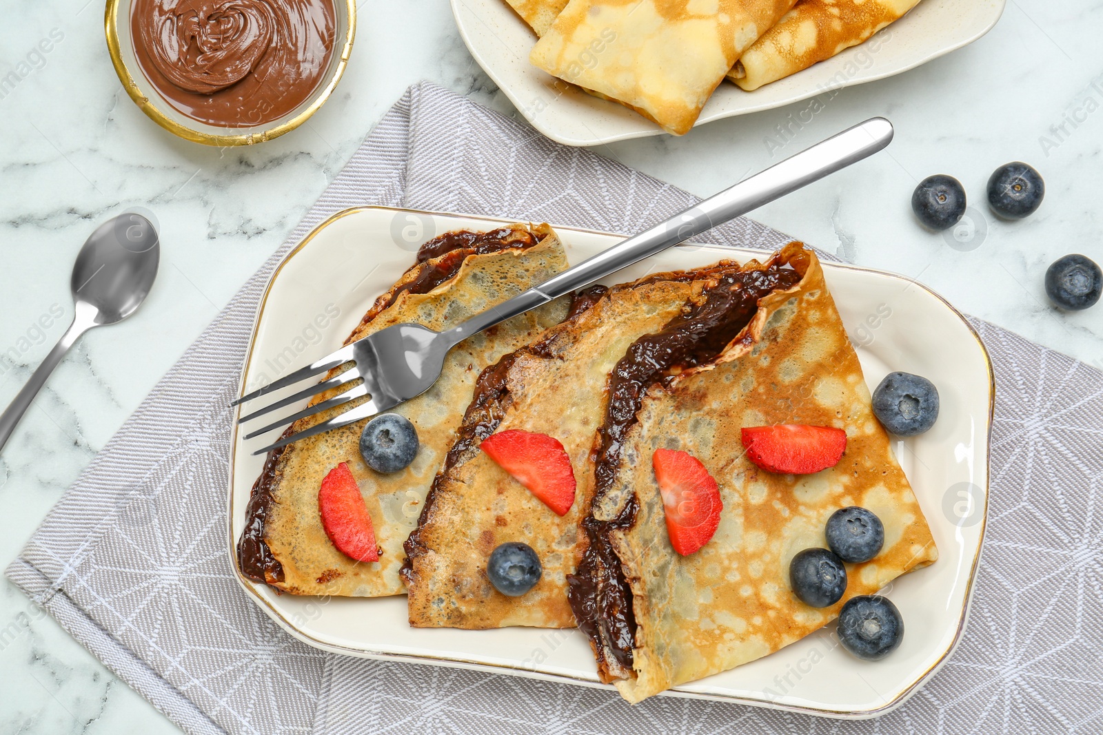 Photo of Tasty crepes with chocolate paste and berries served on white marble table, flat lay