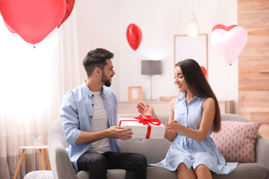 Photo of Young man presenting gift to his girlfriend in living room decorated with heart shaped balloons. Valentine's day celebration
