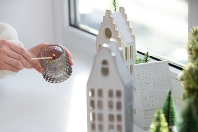 Photo of Woman lightning up candle near windowsill with house shaped lanterns indoors, closeup