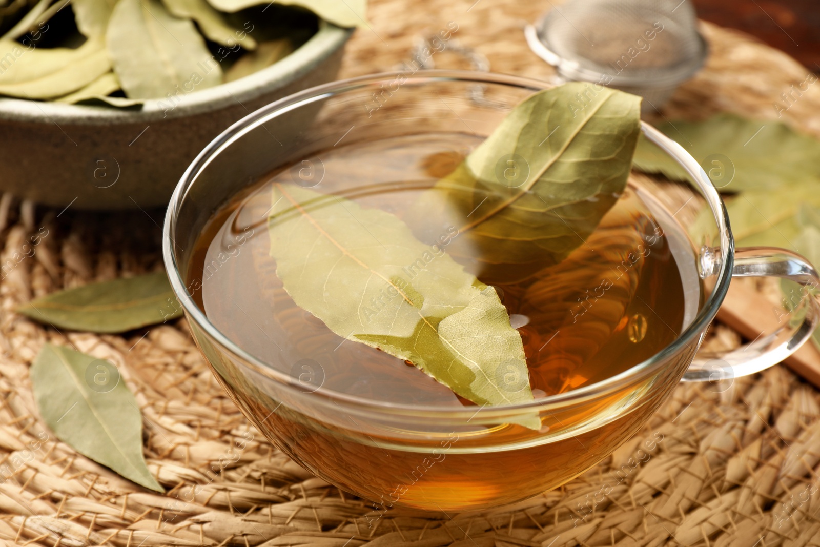 Photo of Cup of freshly brewed tea with bay leaves on wicker mat, closeup