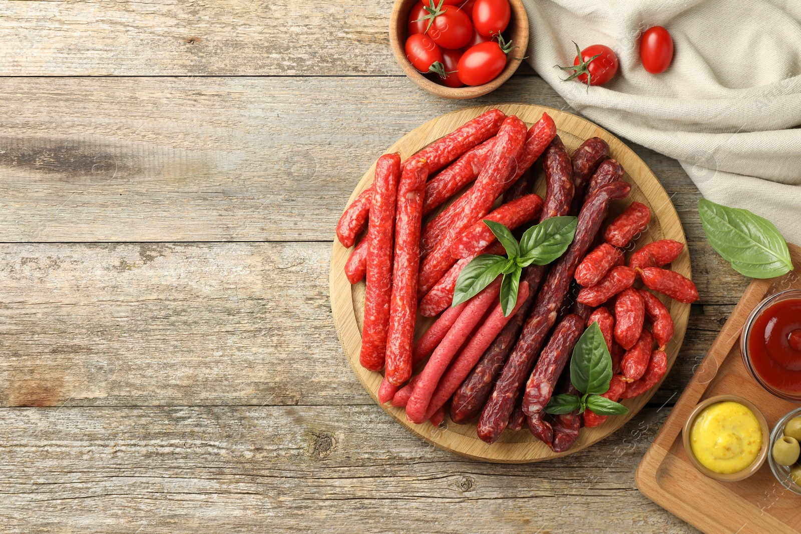 Photo of Different thin dry smoked sausages, basil and sauces on wooden table, flat lay. Space for text