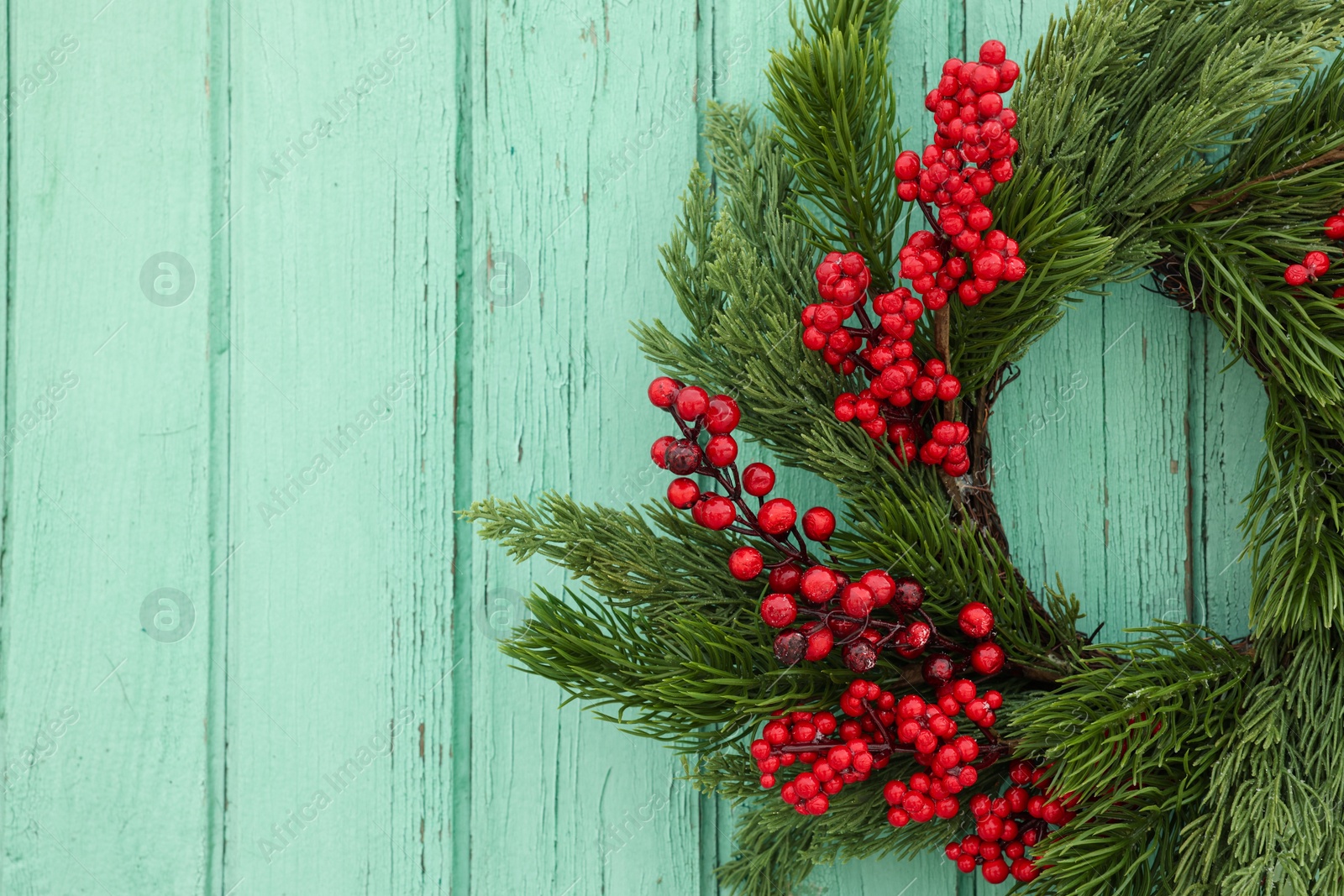 Photo of Beautiful Christmas wreath with red berries hanging on turquoise wooden wall, closeup. Space for text