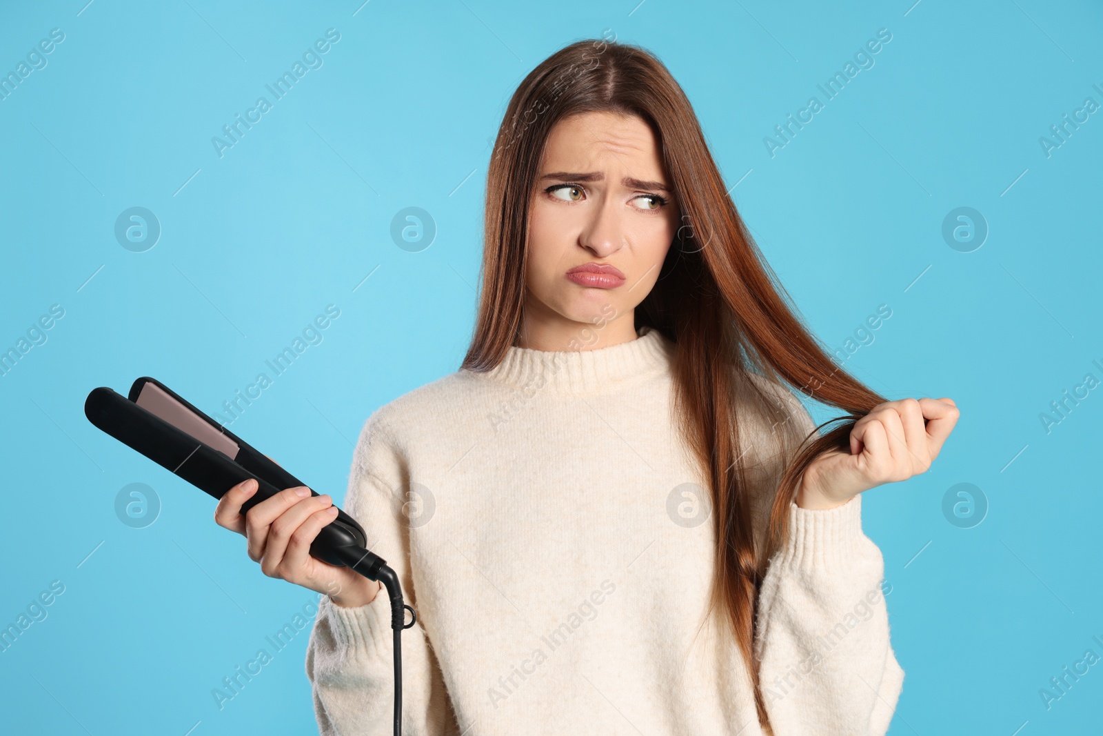 Photo of Upset young woman with flattening iron on light blue background. Hair damage