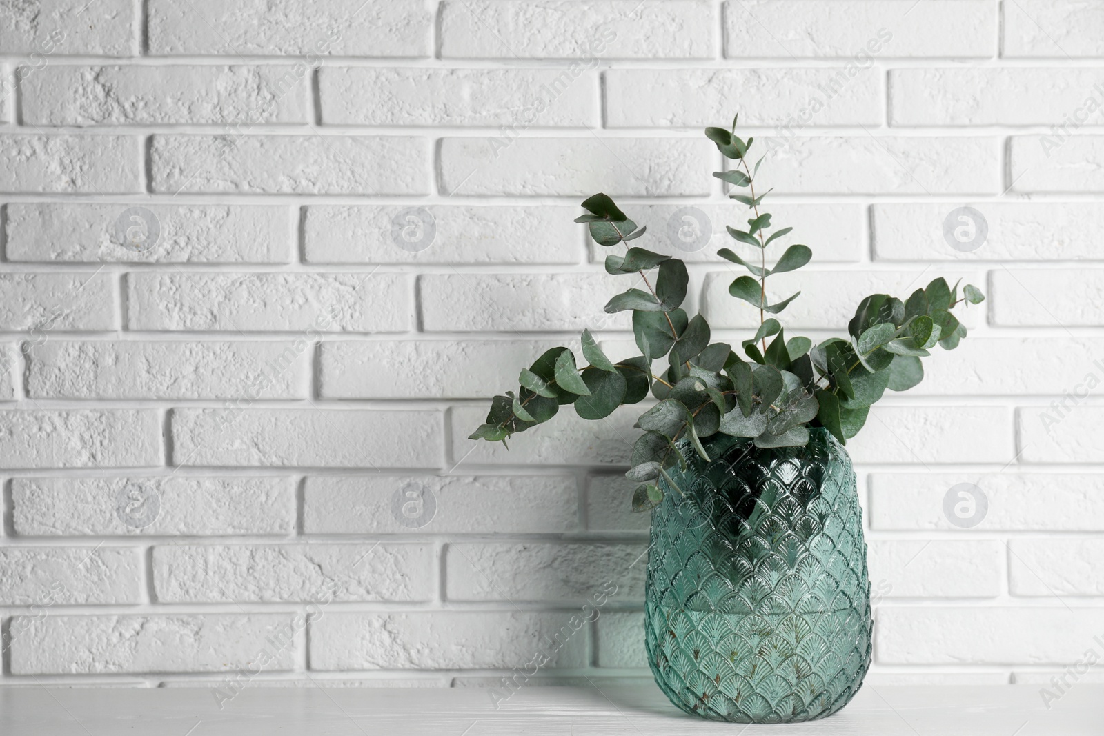 Photo of Beautiful eucalyptus branches in glass vase on white wooden table near brick wall. Space for text