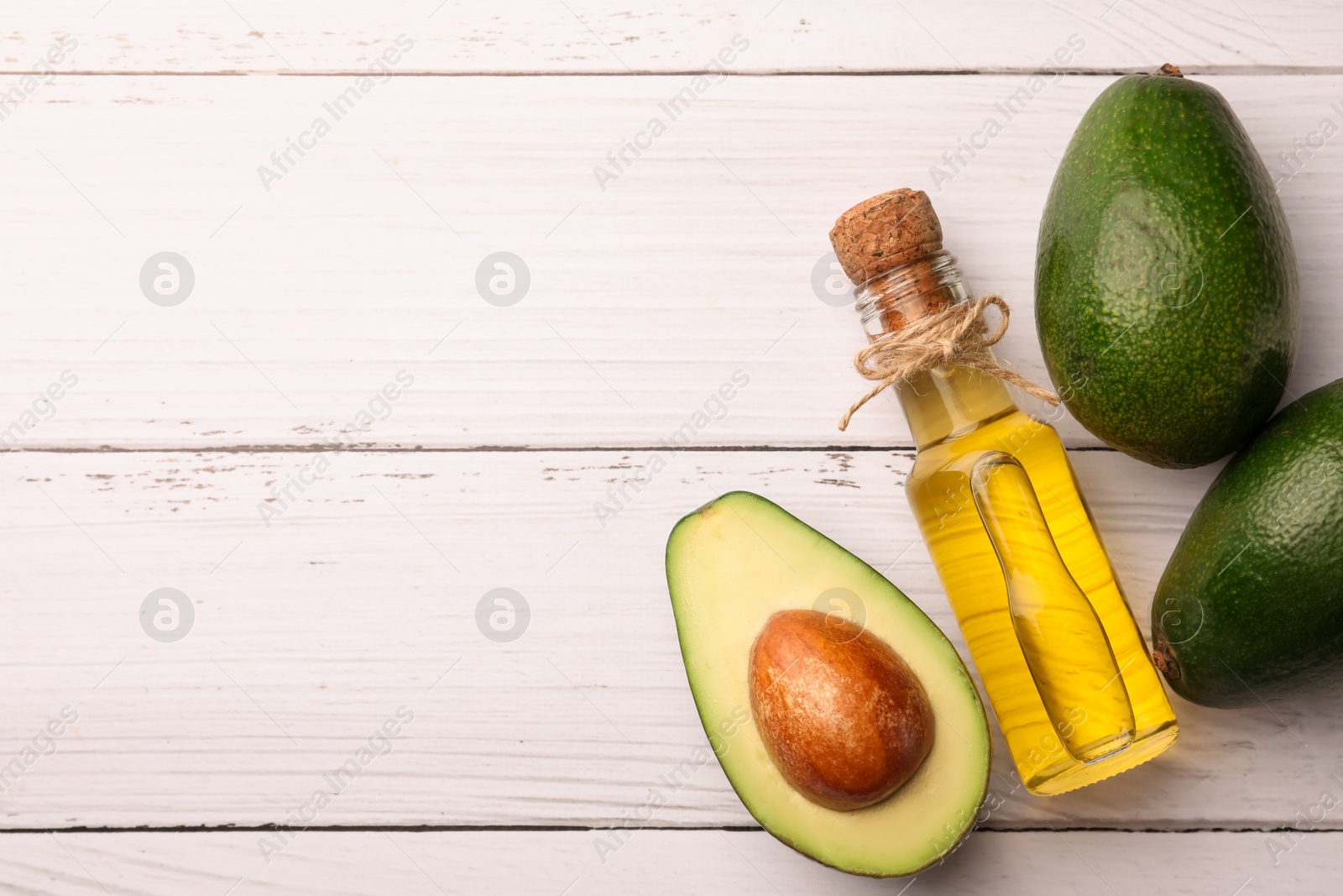 Photo of Glass bottle of cooking oil and fresh avocados on white wooden table, flat lay. Space for text