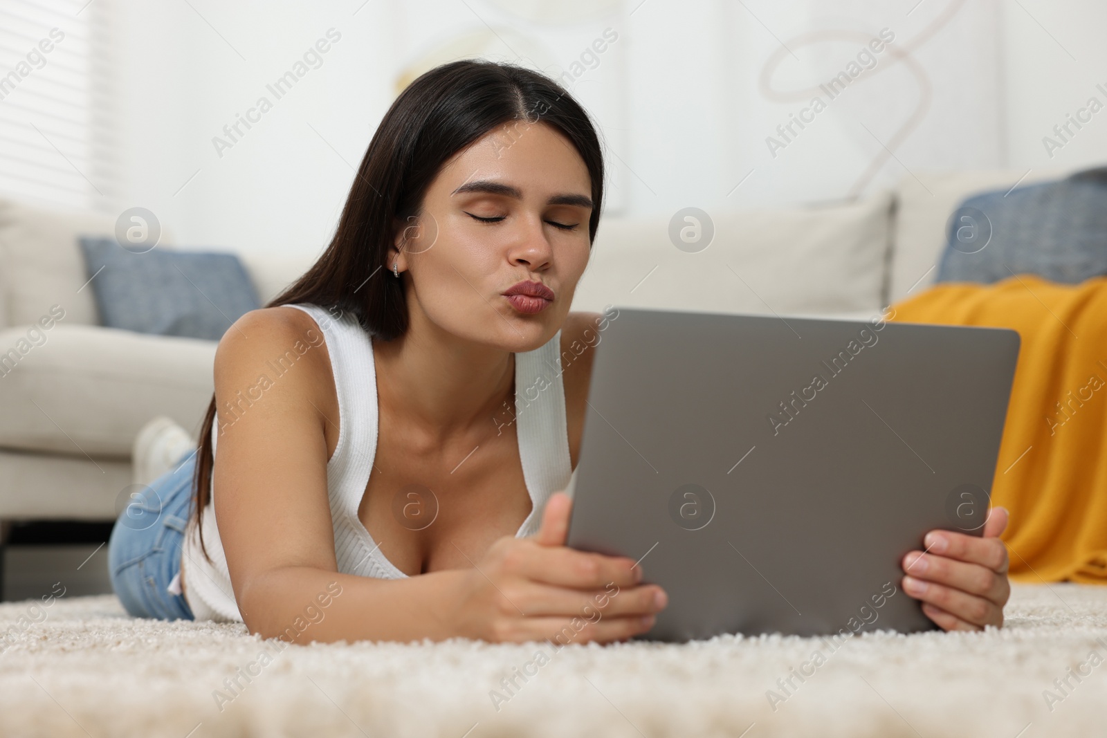 Photo of Young woman having video chat via laptop and blowing kiss on floor at home