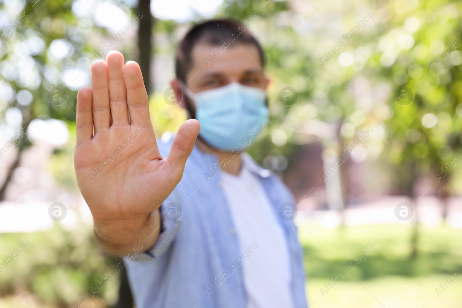 Photo of Man in protective face mask showing stop gesture in park, focus on hand. Prevent spreading of coronavirus