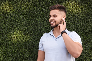 Photo of Young man with wireless headphones listening to music near green grass wall. Space for text