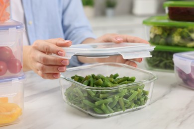 Photo of Woman sealing container with green beans at white marble table in kitchen, closeup. Food storage