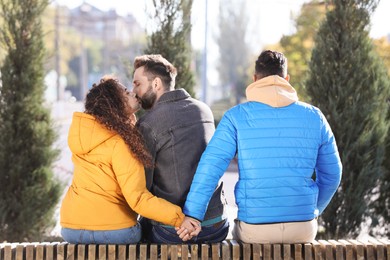 Photo of Woman holding hands with another man while kissing her boyfriend on bench in park. Love triangle