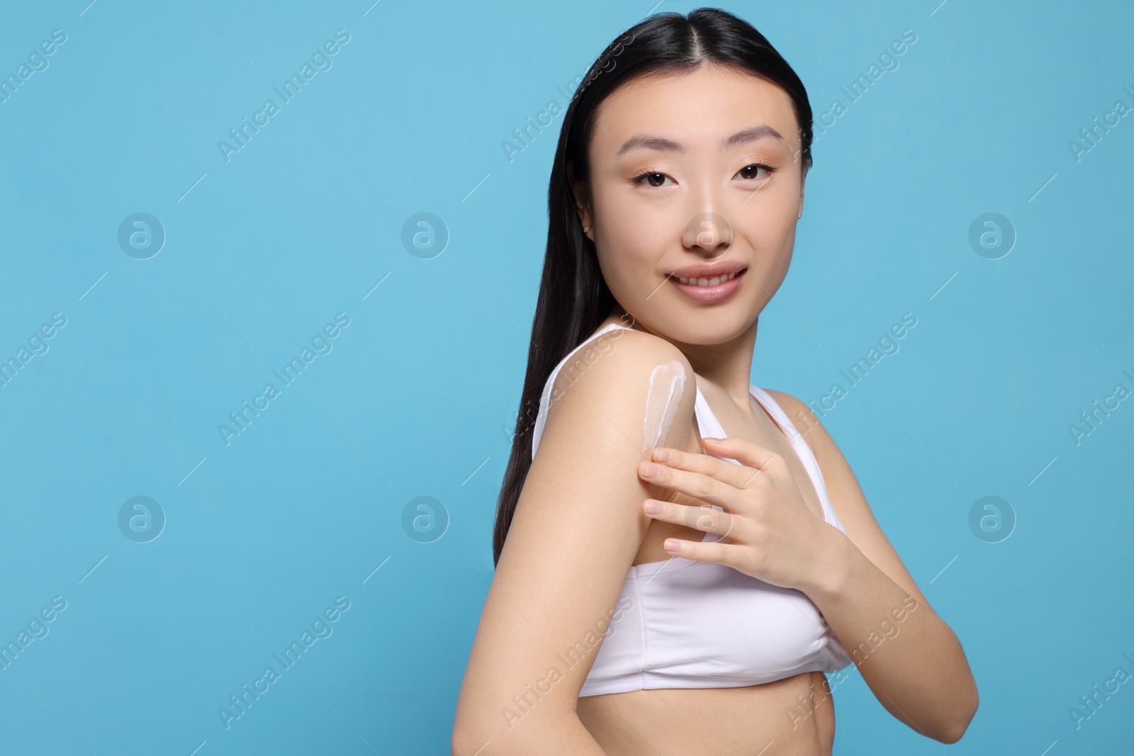 Photo of Beautiful young Asian woman applying body cream onto shoulder on light blue background, space for text