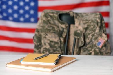 Photo of Notebooks and pen on white wooden table, chair with soldier uniform against flag of United States indoors. Military education