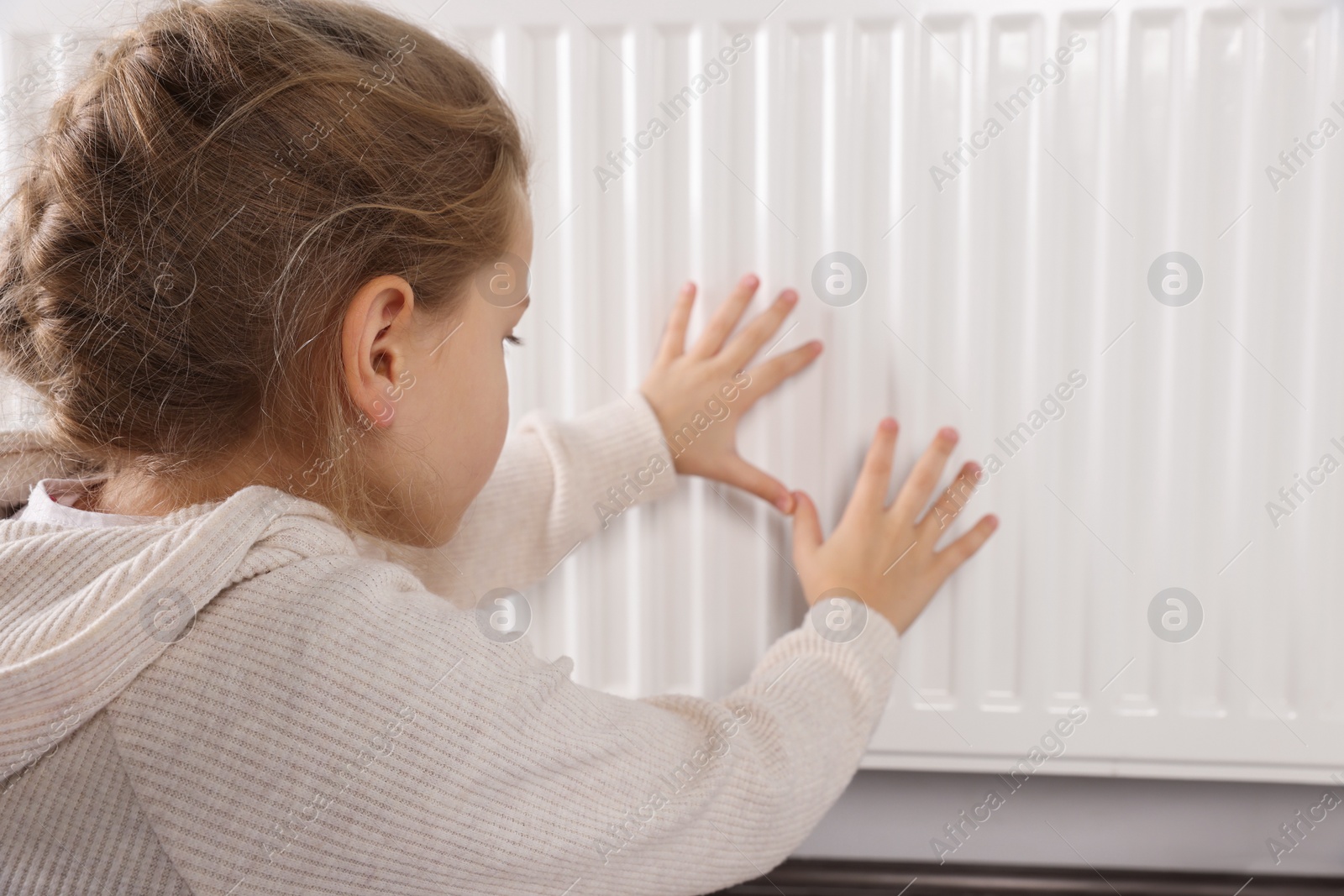 Photo of Little girl warming hands near heating radiator indoors