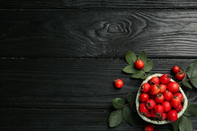 Photo of Ripe rose hip berries with green leaves on black wooden table, flat lay. Space for text