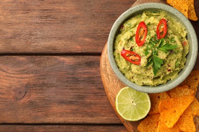 Photo of Bowl of delicious guacamole, lime and nachos chips on wooden table, top view. Space for text