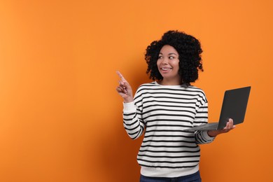 Happy young woman with laptop pointing at something on orange background. Space for text