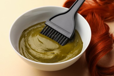 Photo of Bowl of henna cream, brush and red strand on beige background, closeup. Natural hair coloring