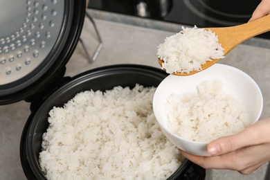 Photo of Woman putting boiled rice into bowl from cooker in kitchen, closeup