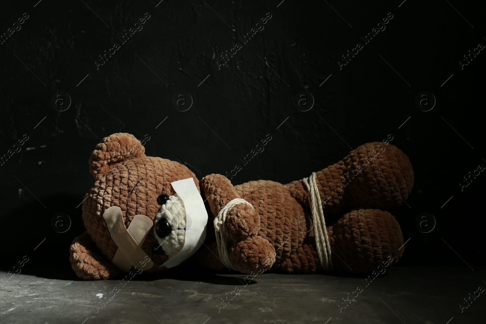 Photo of Stop child abuse. Tied toy bear with taped mouth and patches lying on grey floor against black background. Space for text