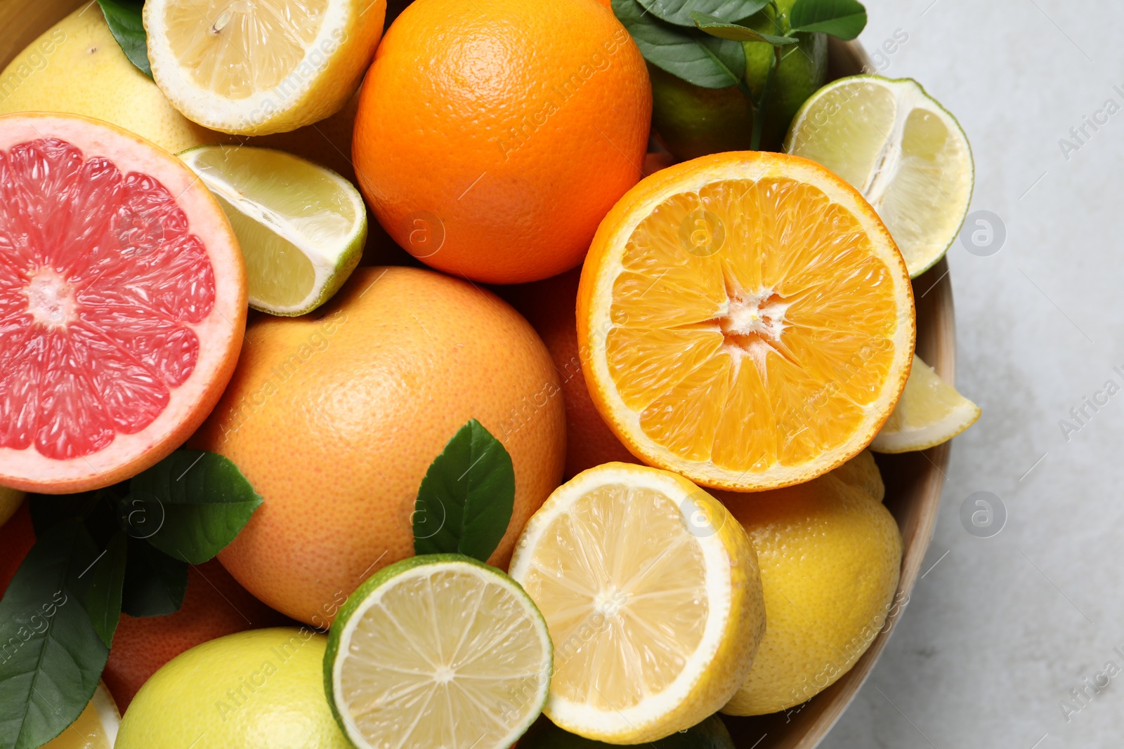 Photo of Different fresh citrus fruits and leaves in bowl on light table, top view