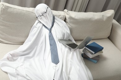 Overworked ghost. Man in white sheet with laptop and books on sofa at home