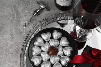 Photo of Bottle, glass of red wine, heart shaped chocolate candies, corkscrew and gift box on light grey textured table, above view. Space for text