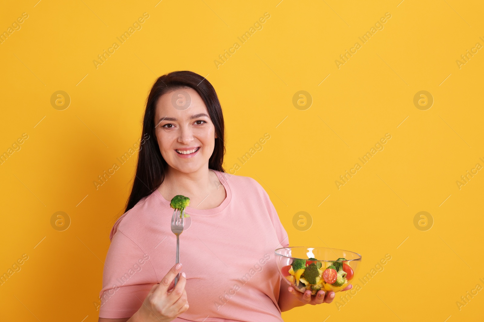 Photo of Beautiful overweight woman eating salad on yellow background, space for text. Healthy diet