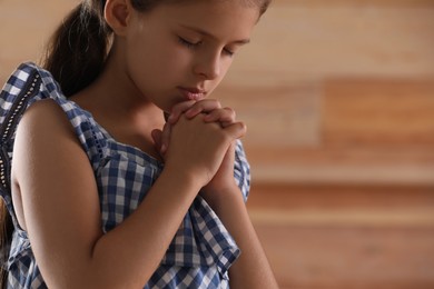 Photo of Cute little girl with hands clasped together praying on blurred background, closeup. Space for text