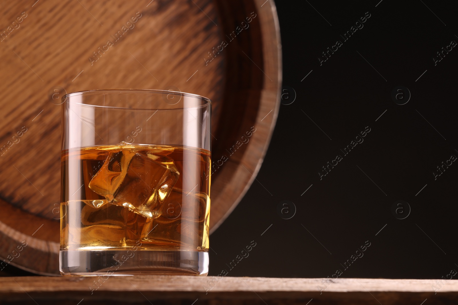 Photo of Whiskey with ice cubes in glass and barrel on wooden table against black background, space for text