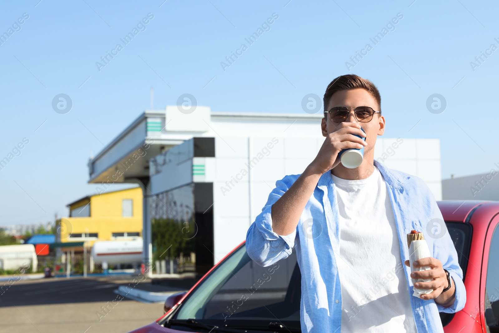 Photo of Young man with hot dog drinking coffee near car at gas station