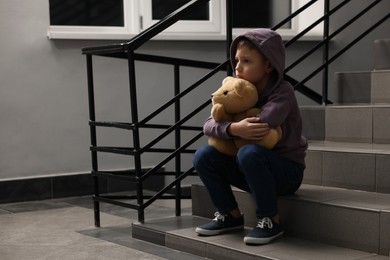 Photo of Child abuse. Upset boy with teddy bear sitting on stairs indoors