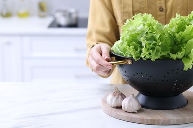 Woman holding black colander with lettuce at white marble table in kitchen, closeup. Space for text