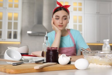 Photo of Upset housewife at messy countertop in kitchen, selective focus. Jar of jam, dishware, eggshells and utensils on table