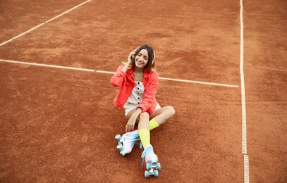 Happy stylish young woman with vintage roller skates and headphones sitting on tennis court