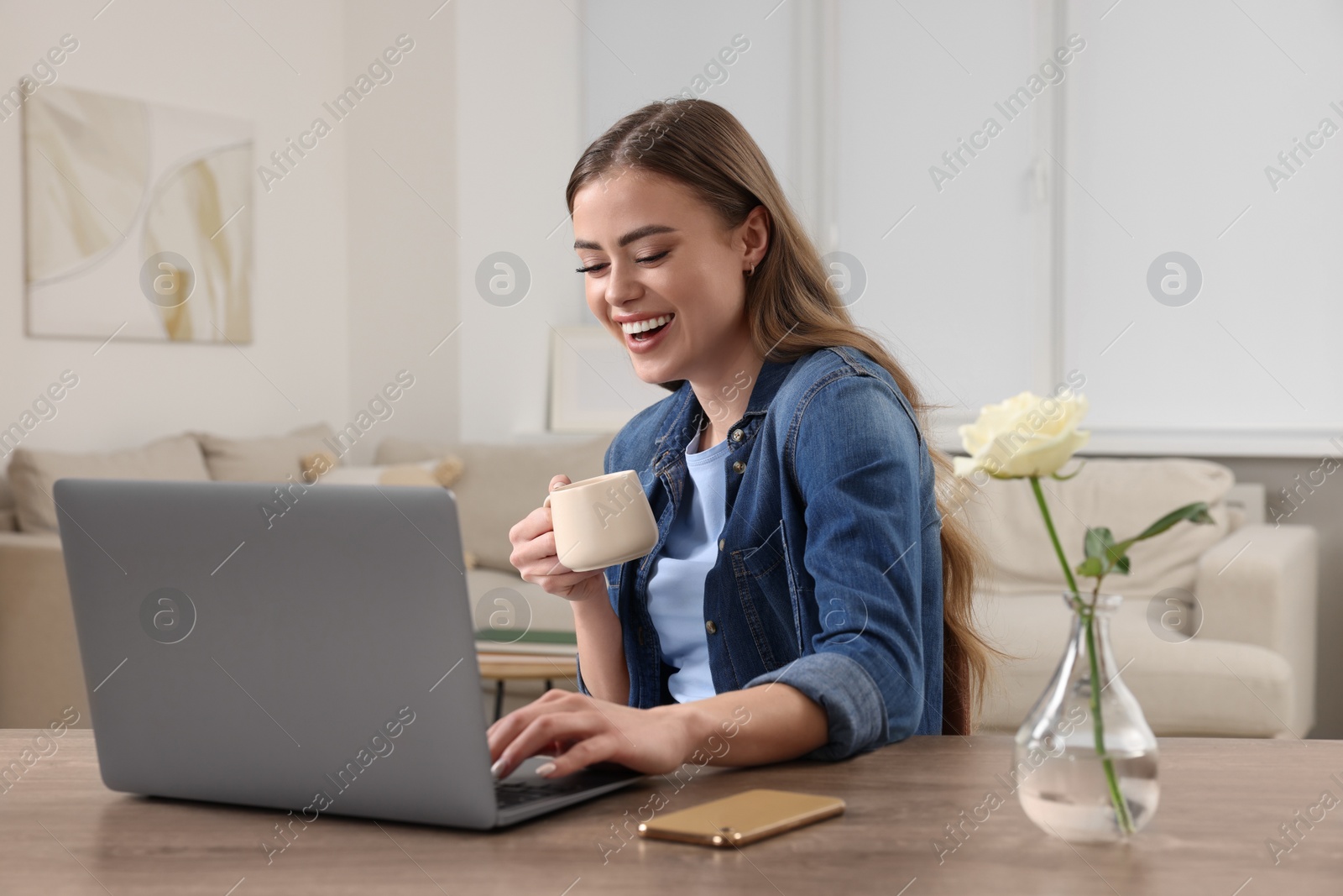 Photo of Happy woman with cup of drink using laptop at wooden table