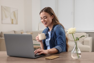 Photo of Happy woman with cup of drink using laptop at wooden table