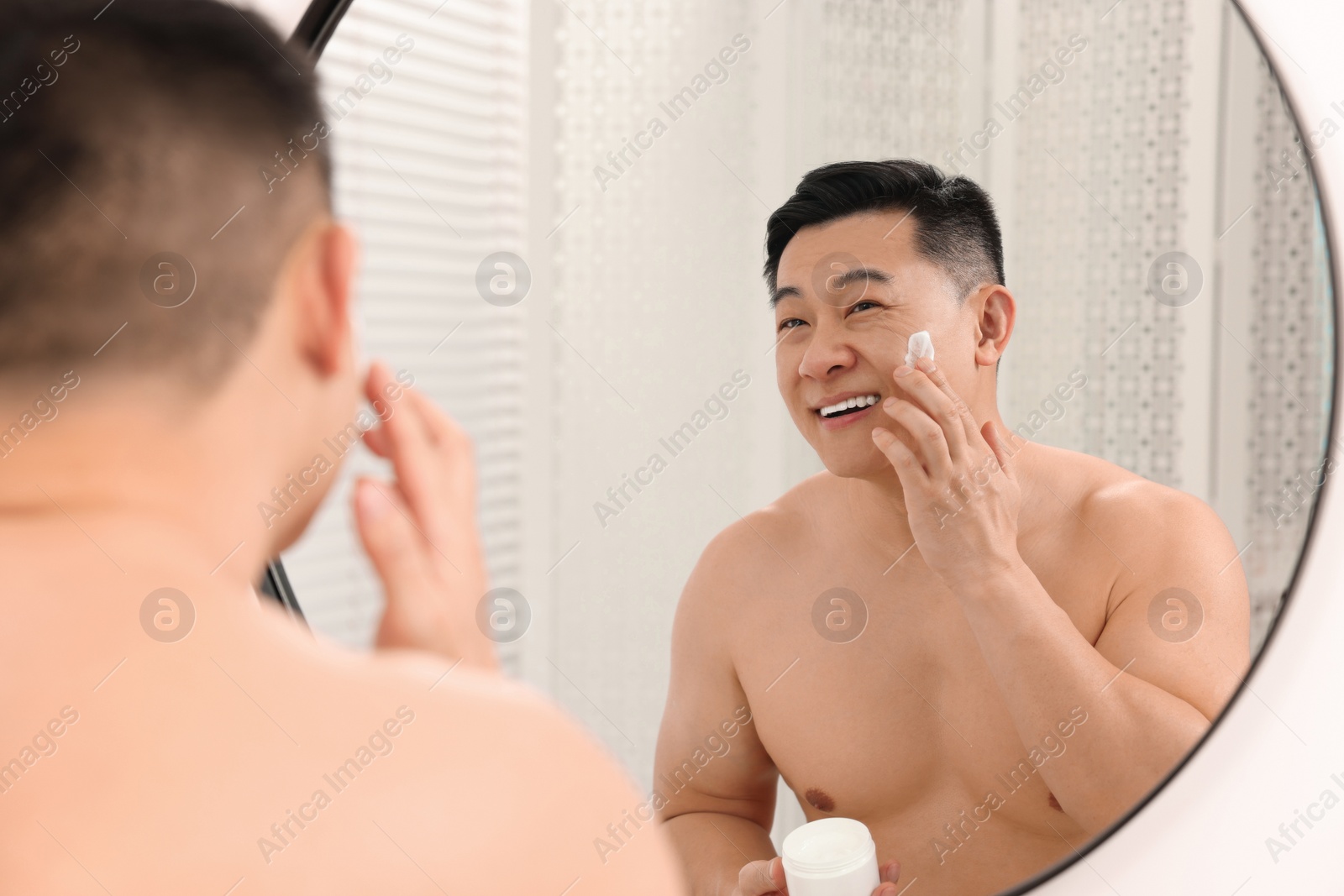 Photo of Handsome man applying cream onto his face near mirror indoors