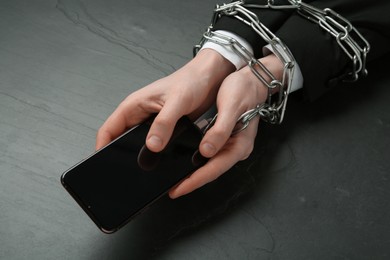 Photo of Man holding smartphone in chained hands at black table, closeup. Internet addiction