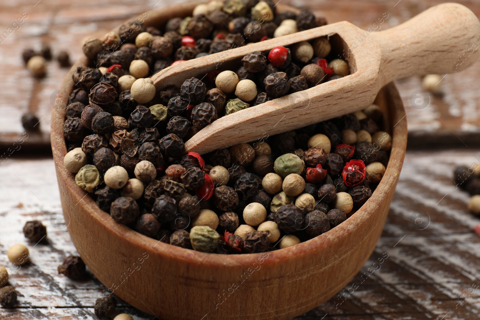 Photo of Aromatic spice. Different peppers in bowl and scoop on wooden table, closeup