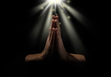 Image of Christian woman holding hands clasped under holy light in darkness, closeup. Prayer and belief