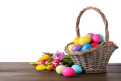 Photo of Colorful Easter eggs in wicker basket and tulips on wooden table against white background