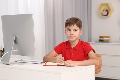 Photo of Boy writing in notepad while using computer at desk in room. Home workplace