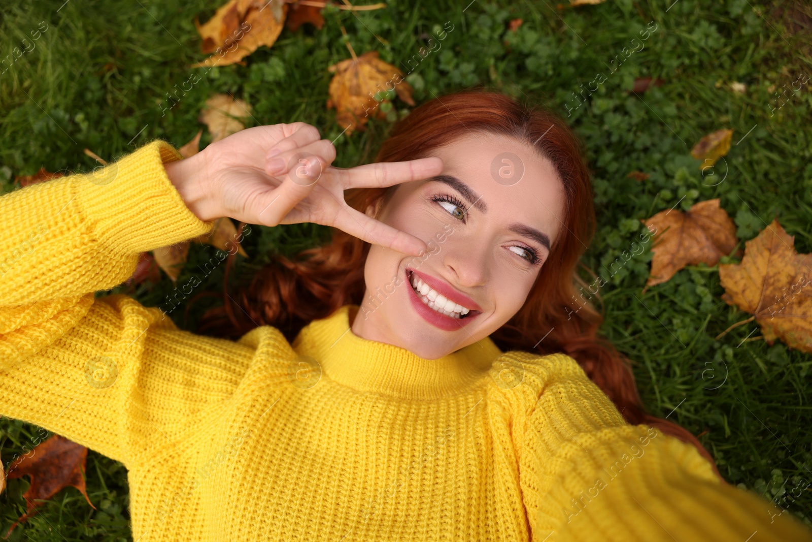 Photo of Smiling woman lying on grass among autumn leaves and taking selfie, top view