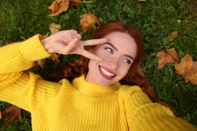Photo of Smiling woman lying on grass among autumn leaves and taking selfie, top view
