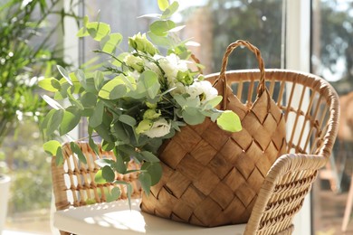 Stylish wicker basket with bouquet of flowers on chair near window indoors